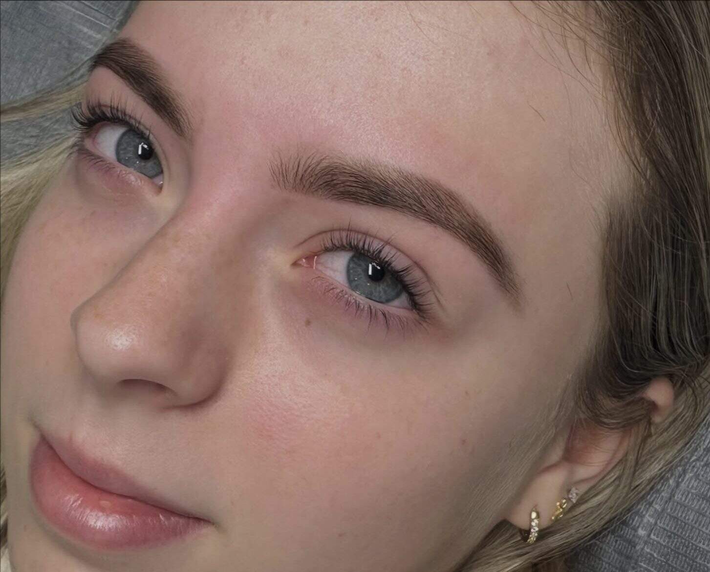 A close-up of a young person with fair skin, blue-gray eyes, well-groomed thick eyebrows, and light blonde hair. The person is wearing small gold earrings and has a calm facial expression.