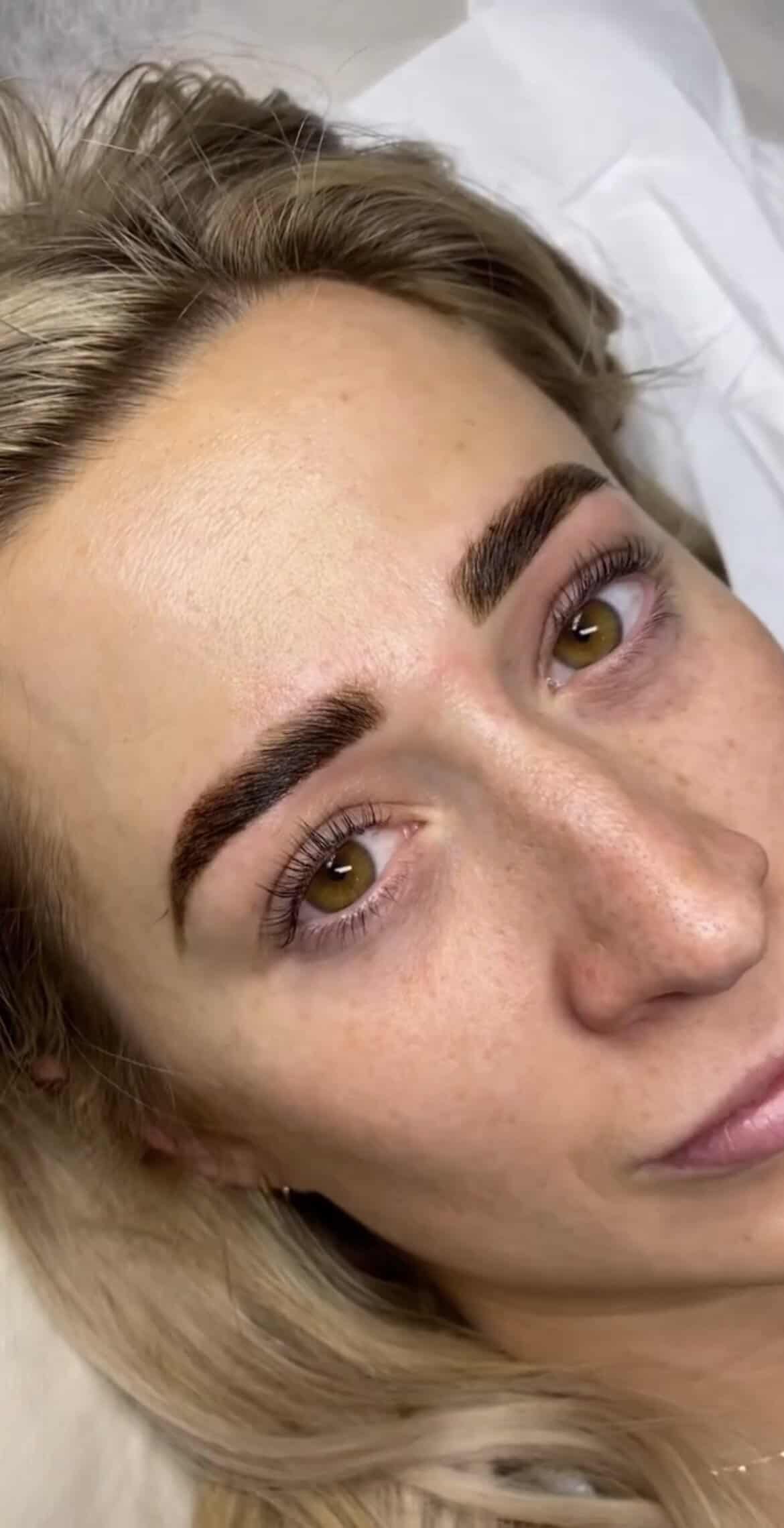 A close-up of a woman with light hair lying down, showing her face and freshly groomed dark eyebrows. Her skin is clear, and she has brown eyes, faint freckles, and a subtle smile.