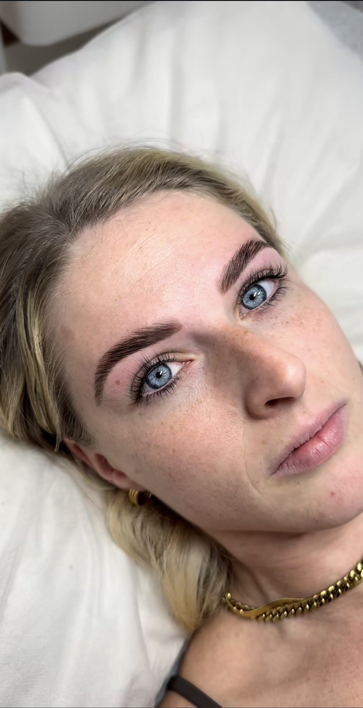 A woman with light skin, blue eyes, and blonde hair lies on a white pillow, looking at the camera. She has well-groomed, defined eyebrows and wears a gold necklace and small gold hoop earrings.