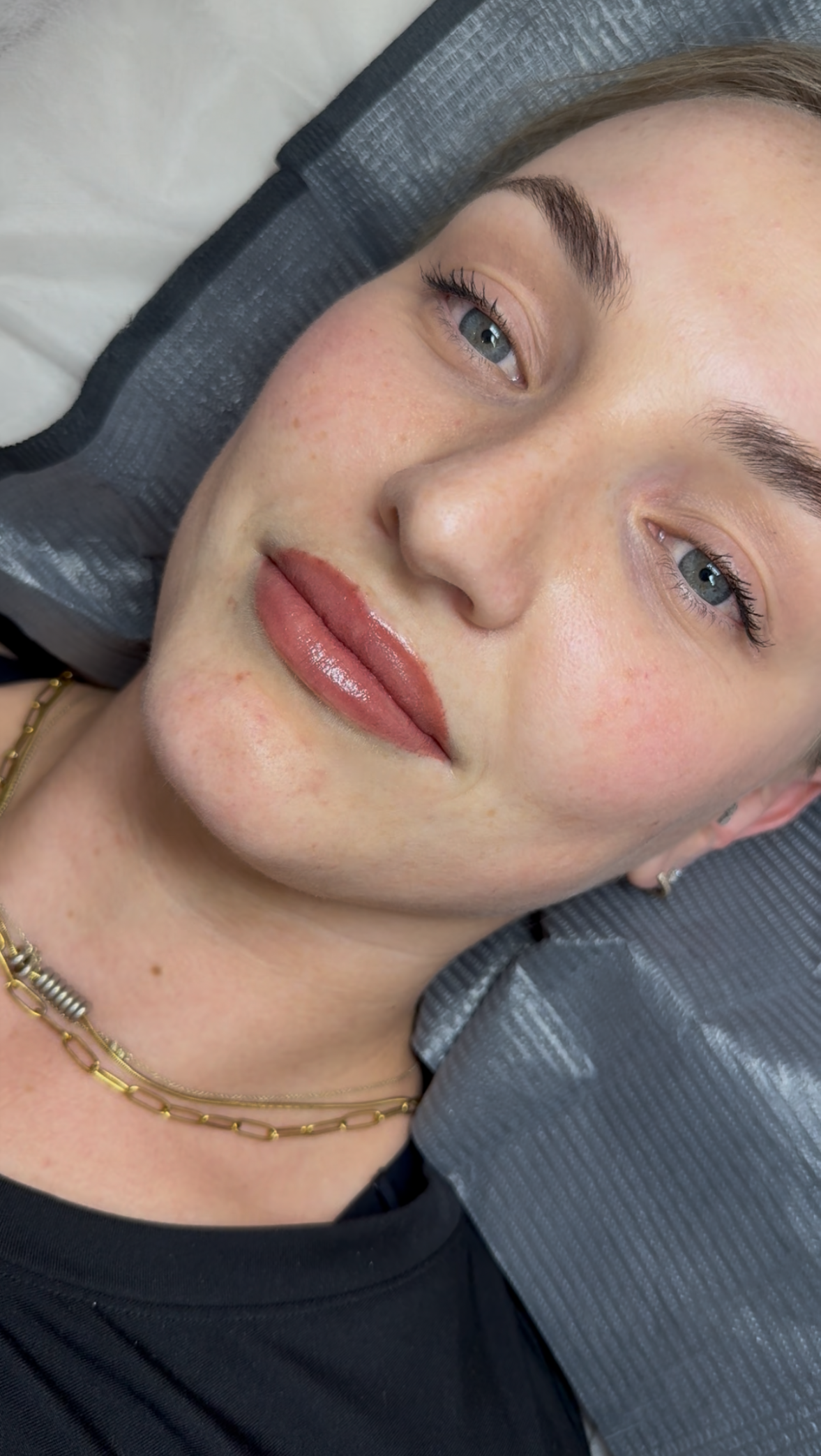 A woman with blue eyes and light skin lies on a gray pillow, wearing a gold chain necklace and a black shirt. She has light makeup, defined brows, and natural-toned lipstick, looking calmly at the camera.