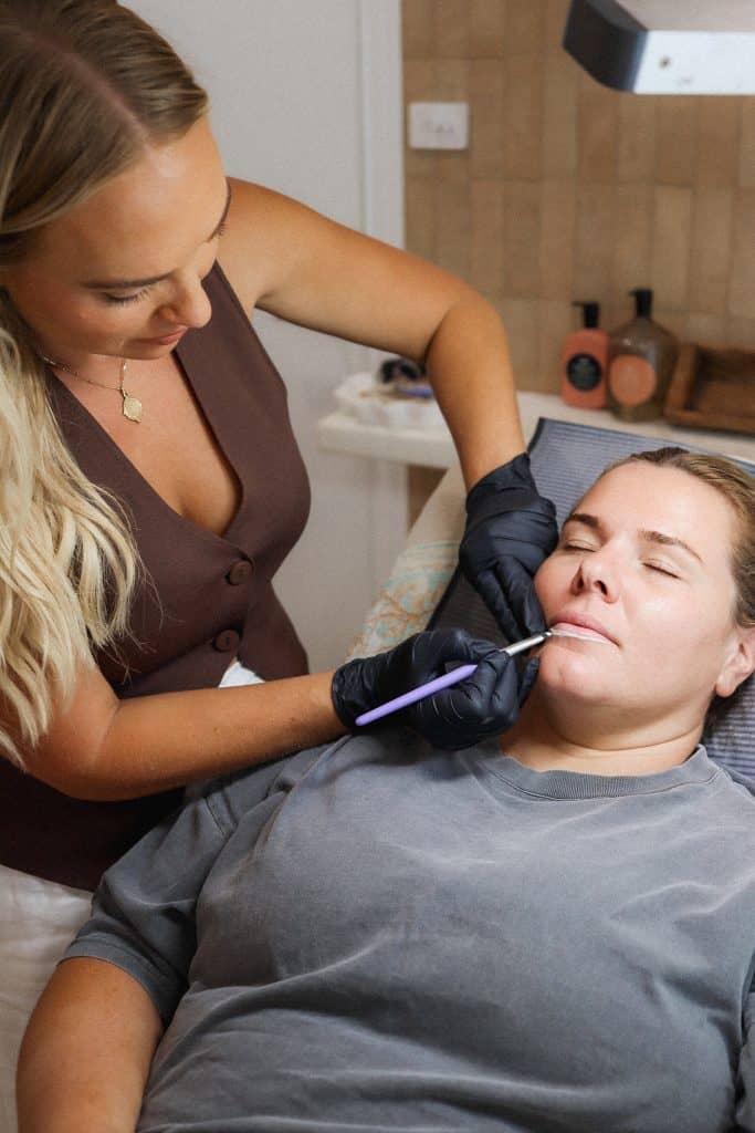 A woman wearing black gloves carefully applies a cosmetic tool to another womans lips, who is lying on a treatment bed in a clinic setting.