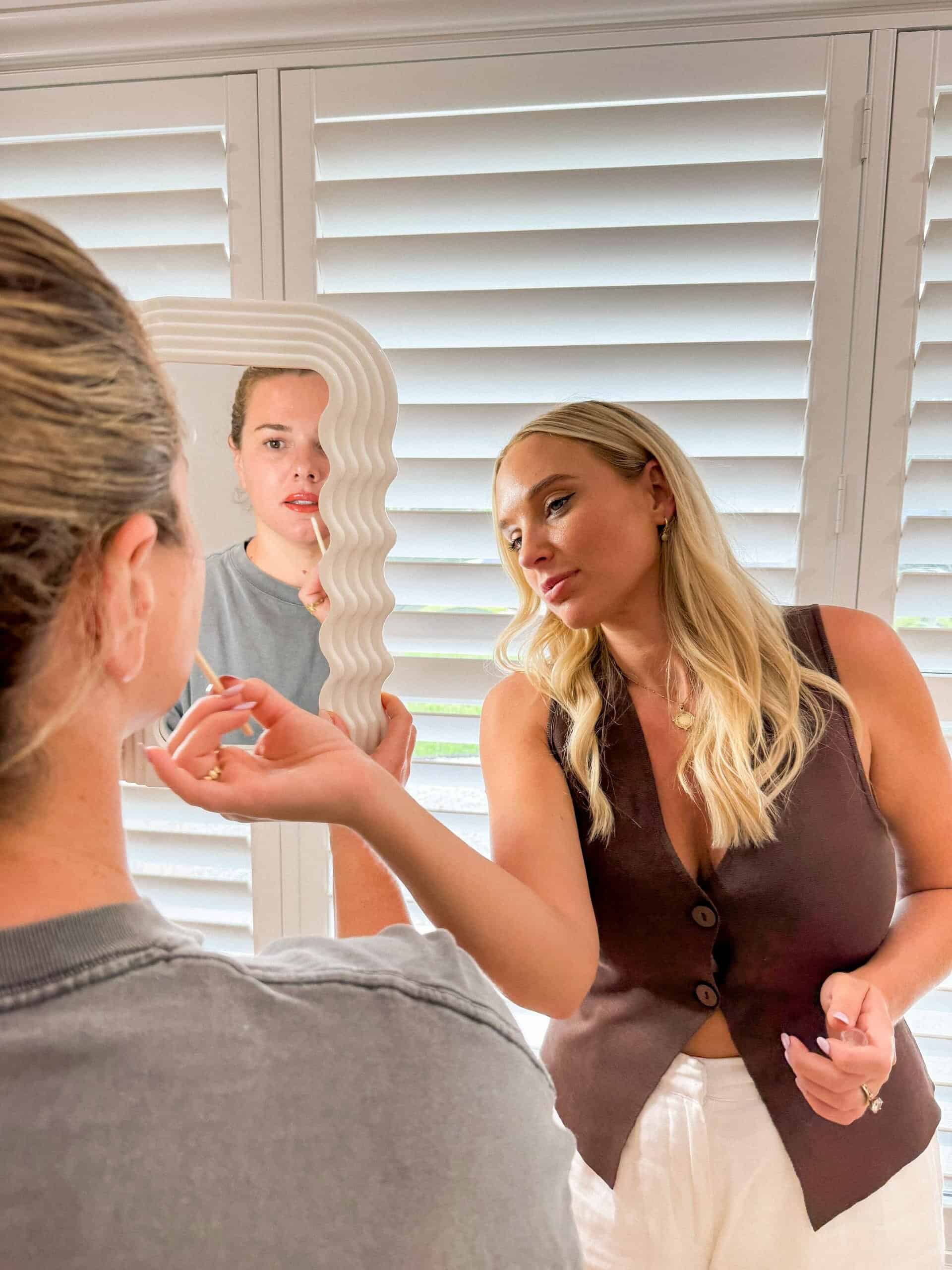 A woman with long blonde hair and a brown vest looks into a wavy-edged mirror held by another person, whose reflection is visible. They stand in front of white window shutters.